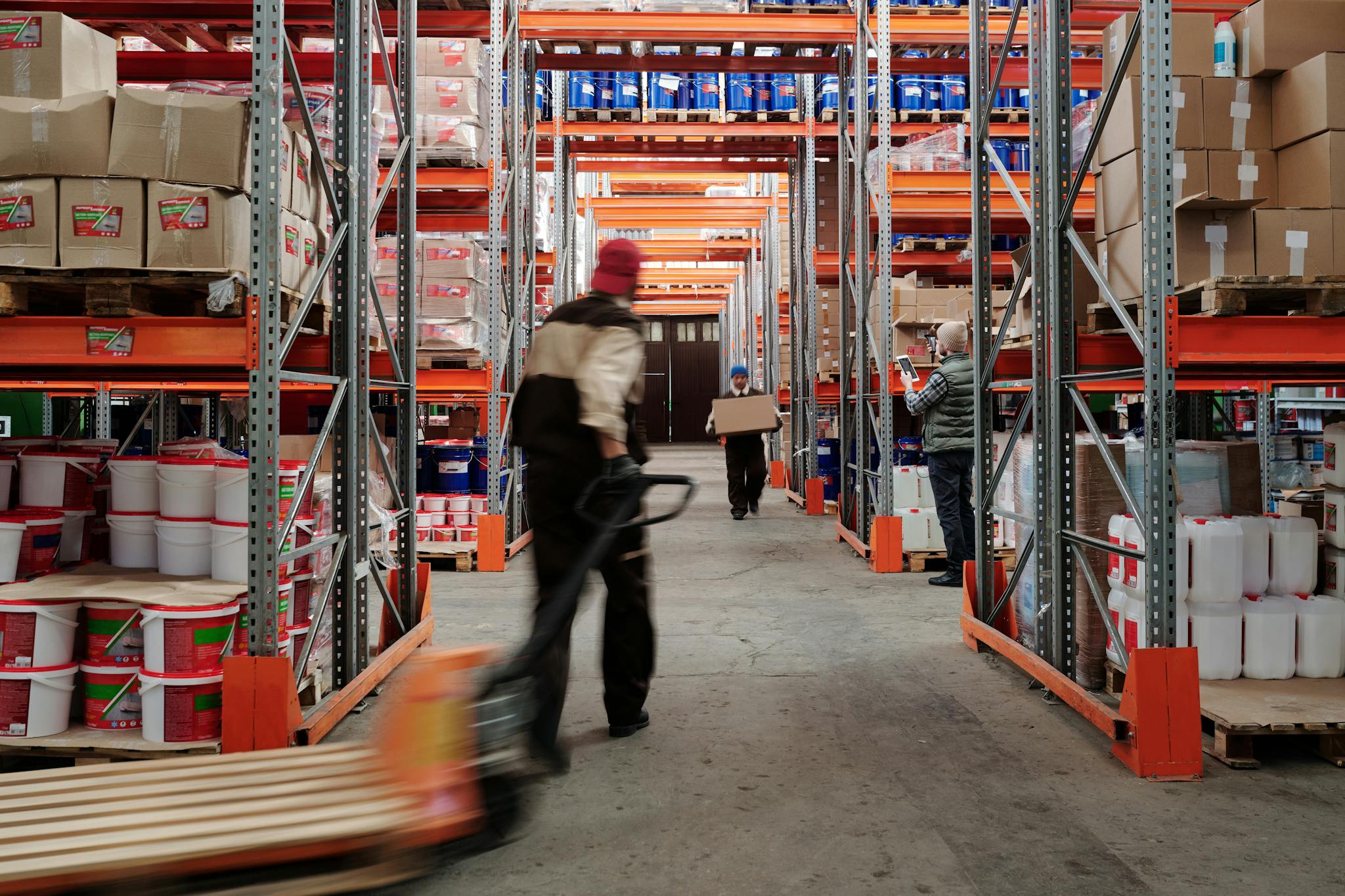Image shows two employees in a warehouse walking towards one another carrying supplies and one is using a dolly.