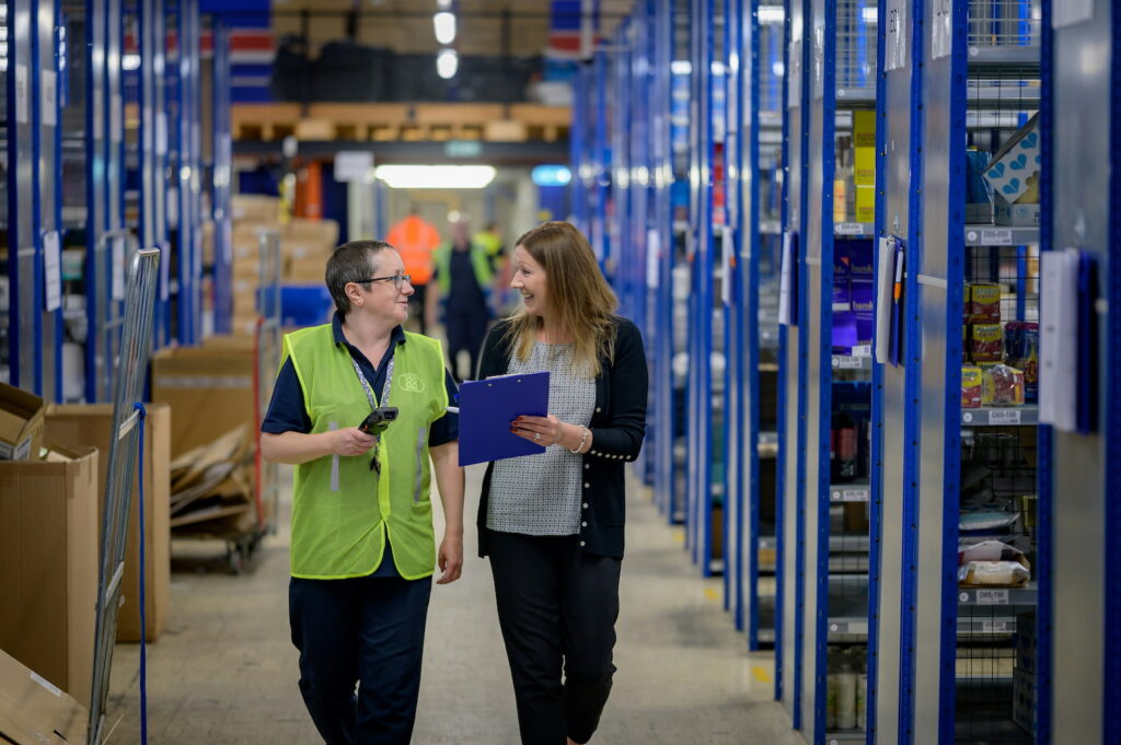 Image of two employees walking down an aisle in a large warehouse, talking in a friendly manner.