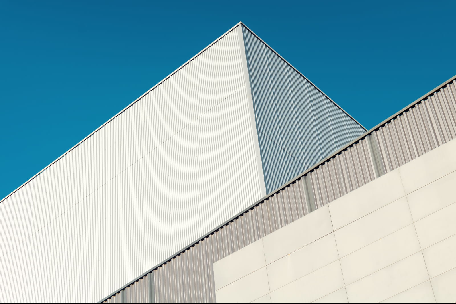 Image is of the tops of steel colored corrugated metal buildings that jut into the blue sky overhead. 