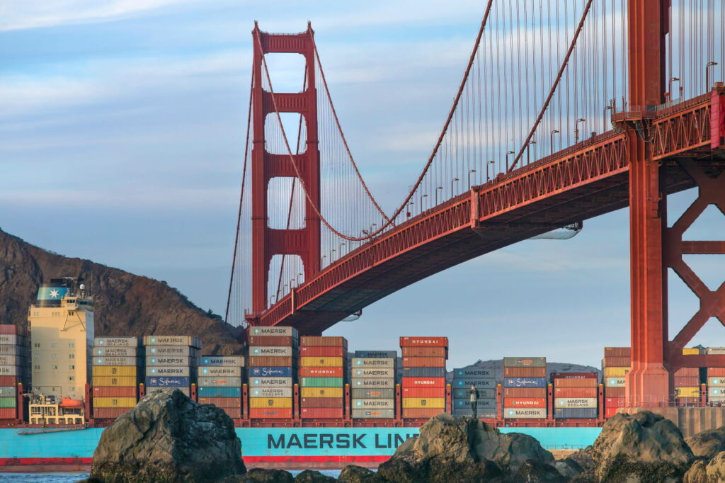 The image shows the San Francisco bridge with a container ship passing slowly underneath, on an overcast day with mountain in background.